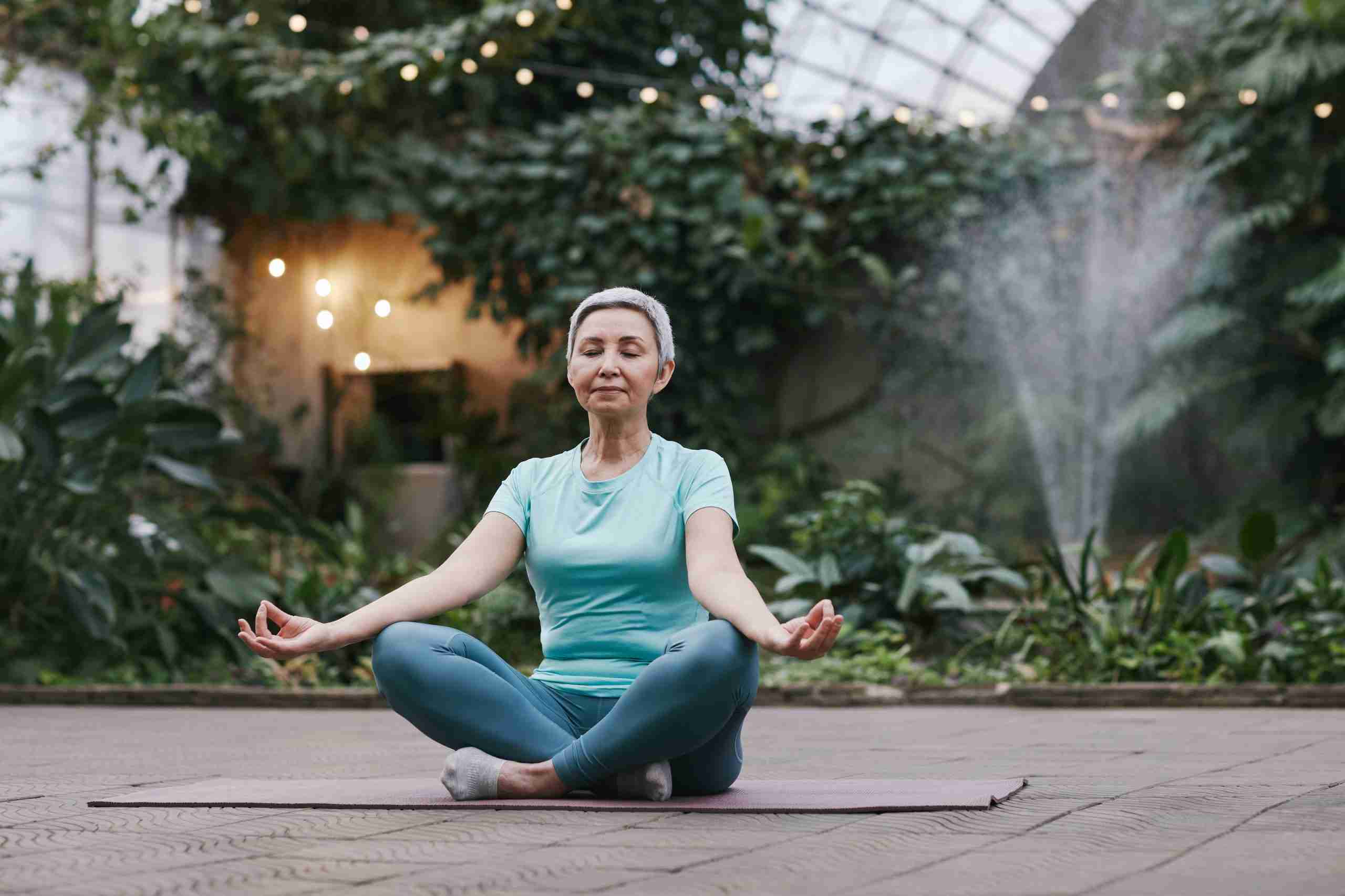 starija dama sjedi na prostirci za jogu meditirajući u zelenom lijepom vrtu https://before-night-falls.com/ zdravlja i ljepote